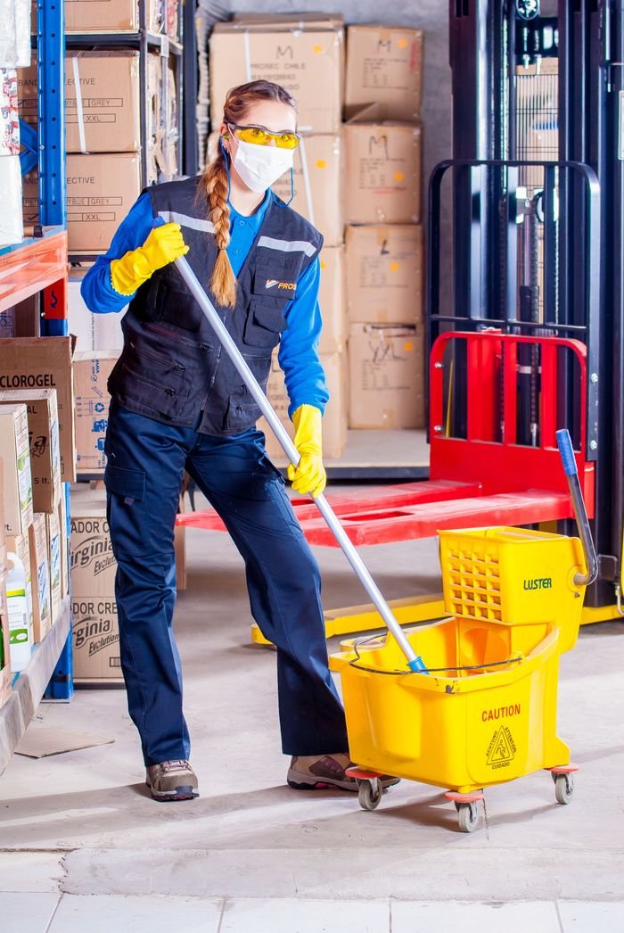 why-choose-us Female janitor in safety gear cleaning a warehouse floor with a mop and bucket.